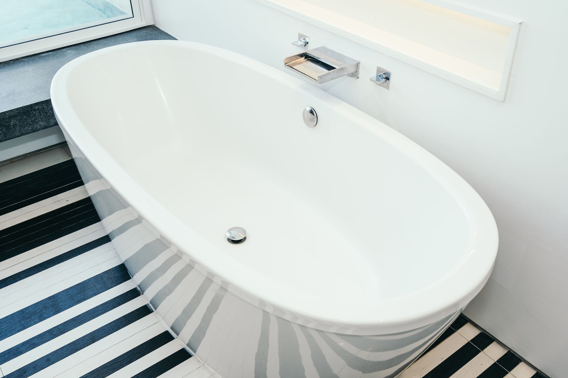 White oval bathtub beside a window in a bright bathroom with black-and-white striped flooring