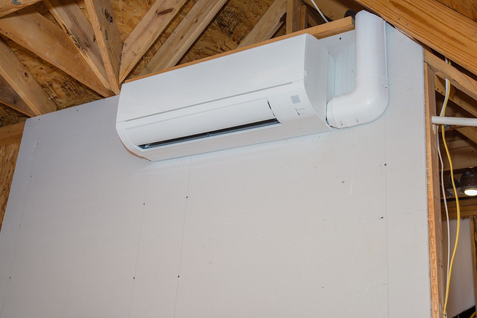 White mini-split air conditioner mounted on a white wall in a room with wood rafters overhead.