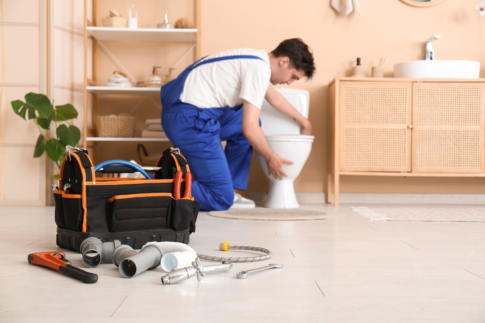 Plumber in blue overalls repairing a toilet in a bathroom with toolbox and tools.