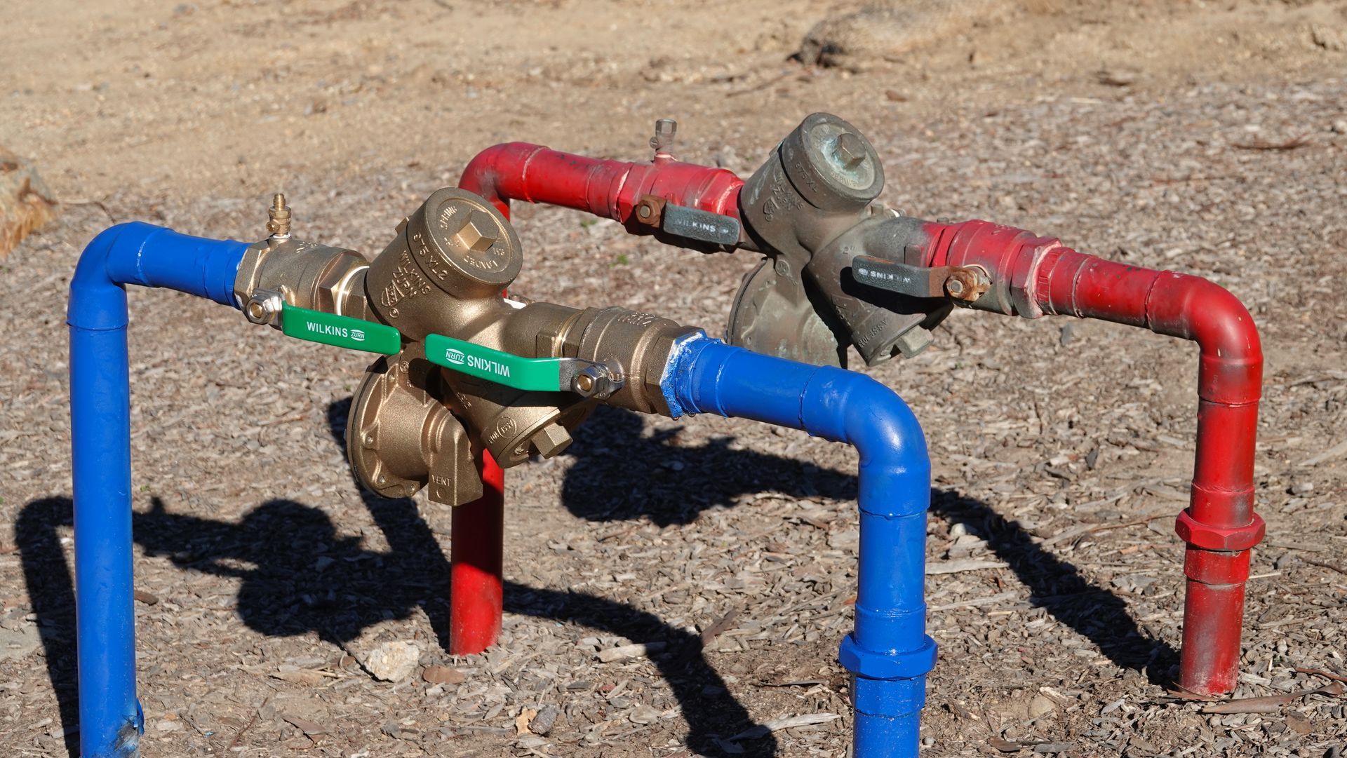 Two parallel pipes, one red, one blue, connected to bronze valves with green handles, outdoors.