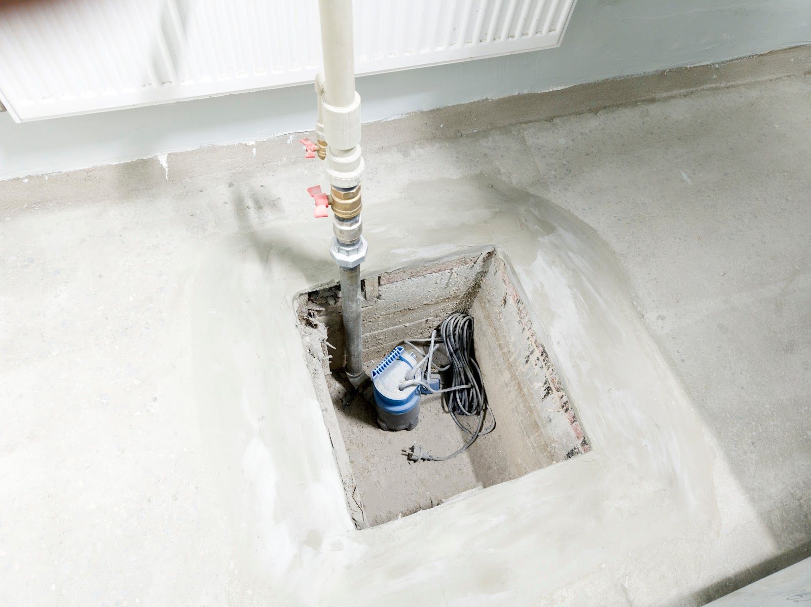 Sump pump installed in concrete floor, with a pipe leading upwards to a valve and heating radiator.
