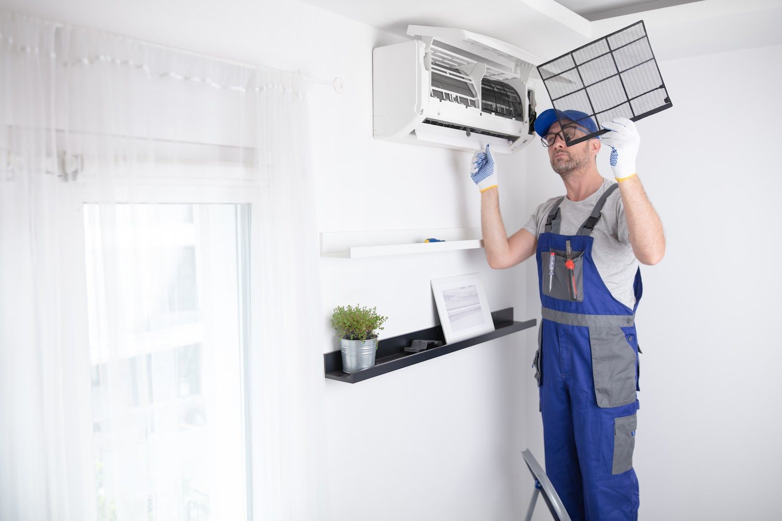 Man in blue overalls cleaning an air conditioner filter indoors.