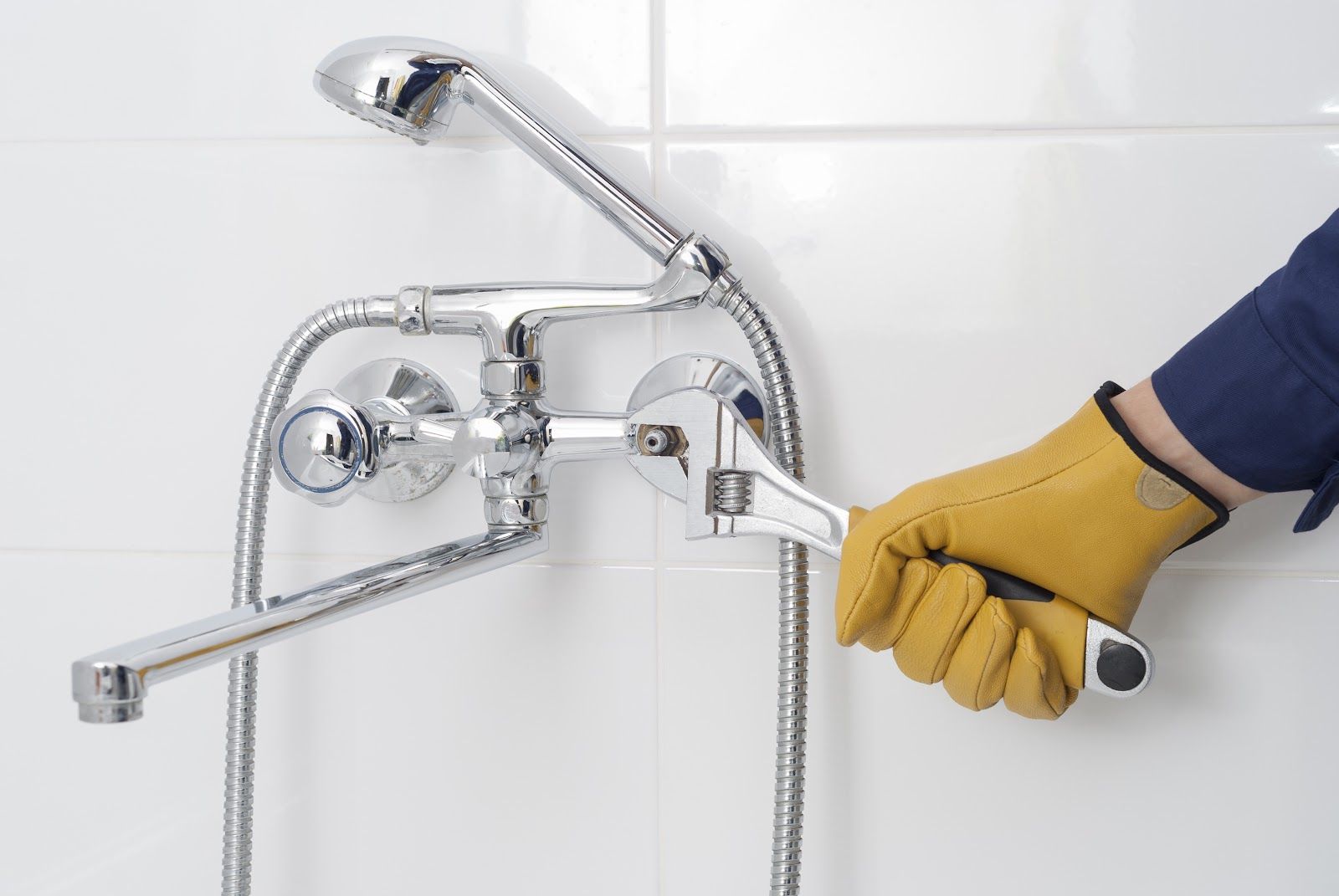 Person in yellow glove uses a wrench to repair a chrome shower fixture against a white-tiled wall.