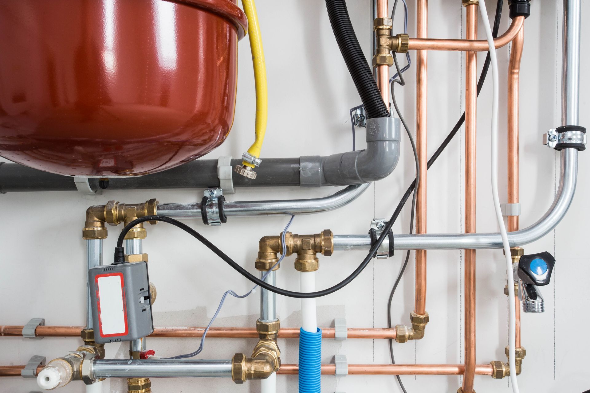 Boiler room piping and valves beneath a red expansion tank, with copper and gray pipes against a white wall