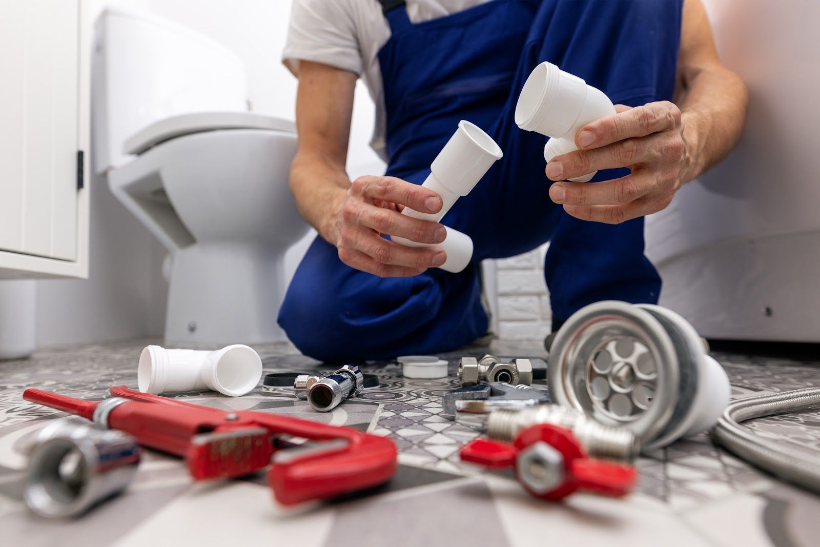 Plumber assembling plumbing parts in a bathroom, toilet in the background, tools and fixtures on the floor.