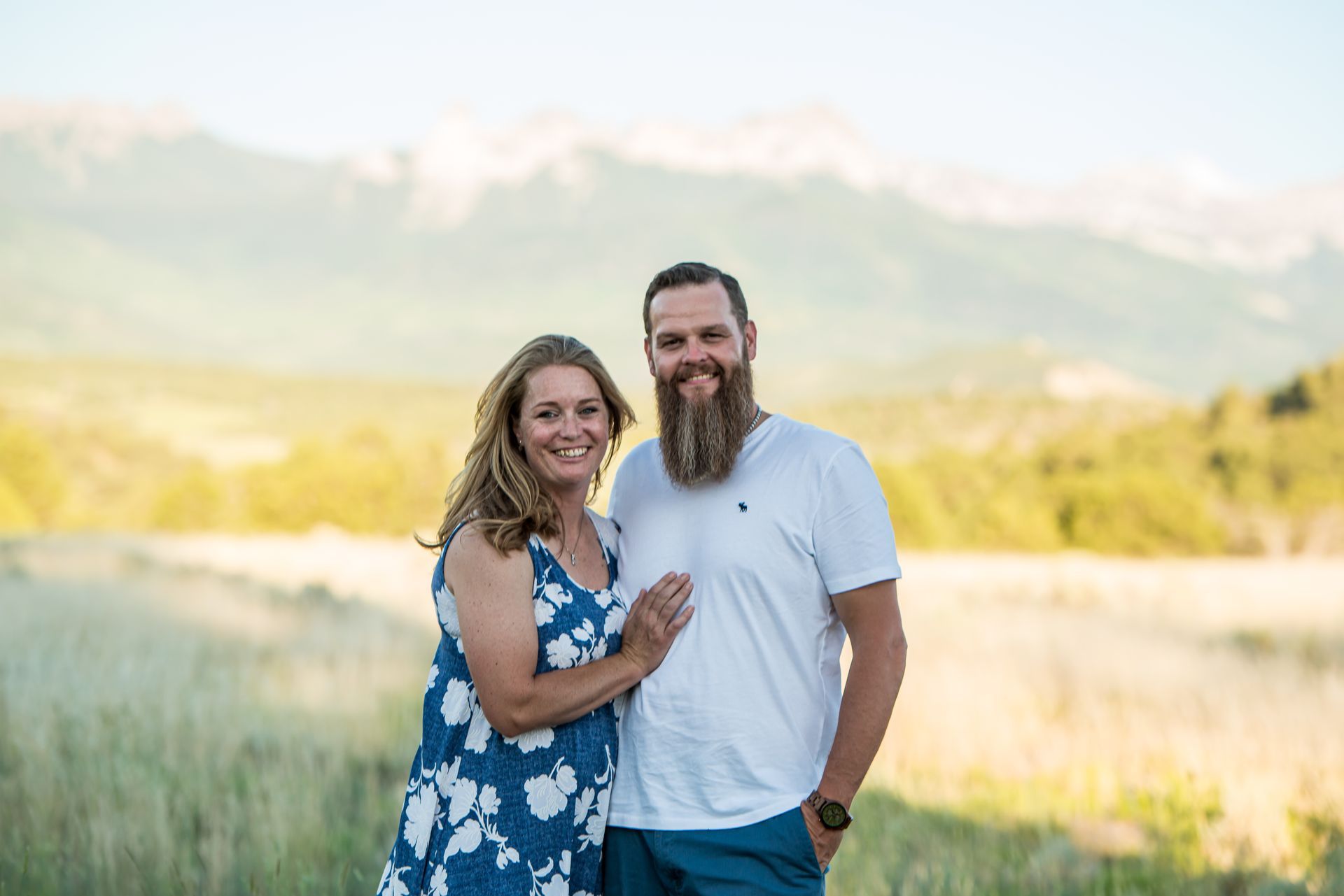 A couple stands in a grassy field before a mountain range, smiling for the camera.