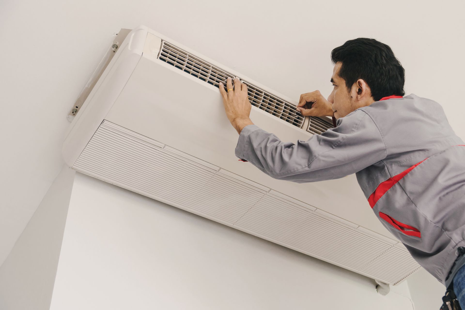Man in gray uniform servicing a white wall-mounted air conditioner.