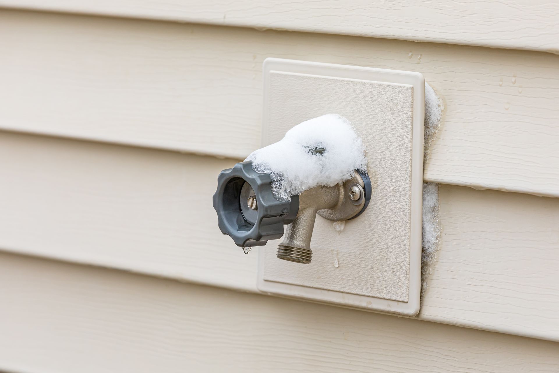 Snow-covered outdoor water spigot on a light beige house, with snow collecting on the siding.