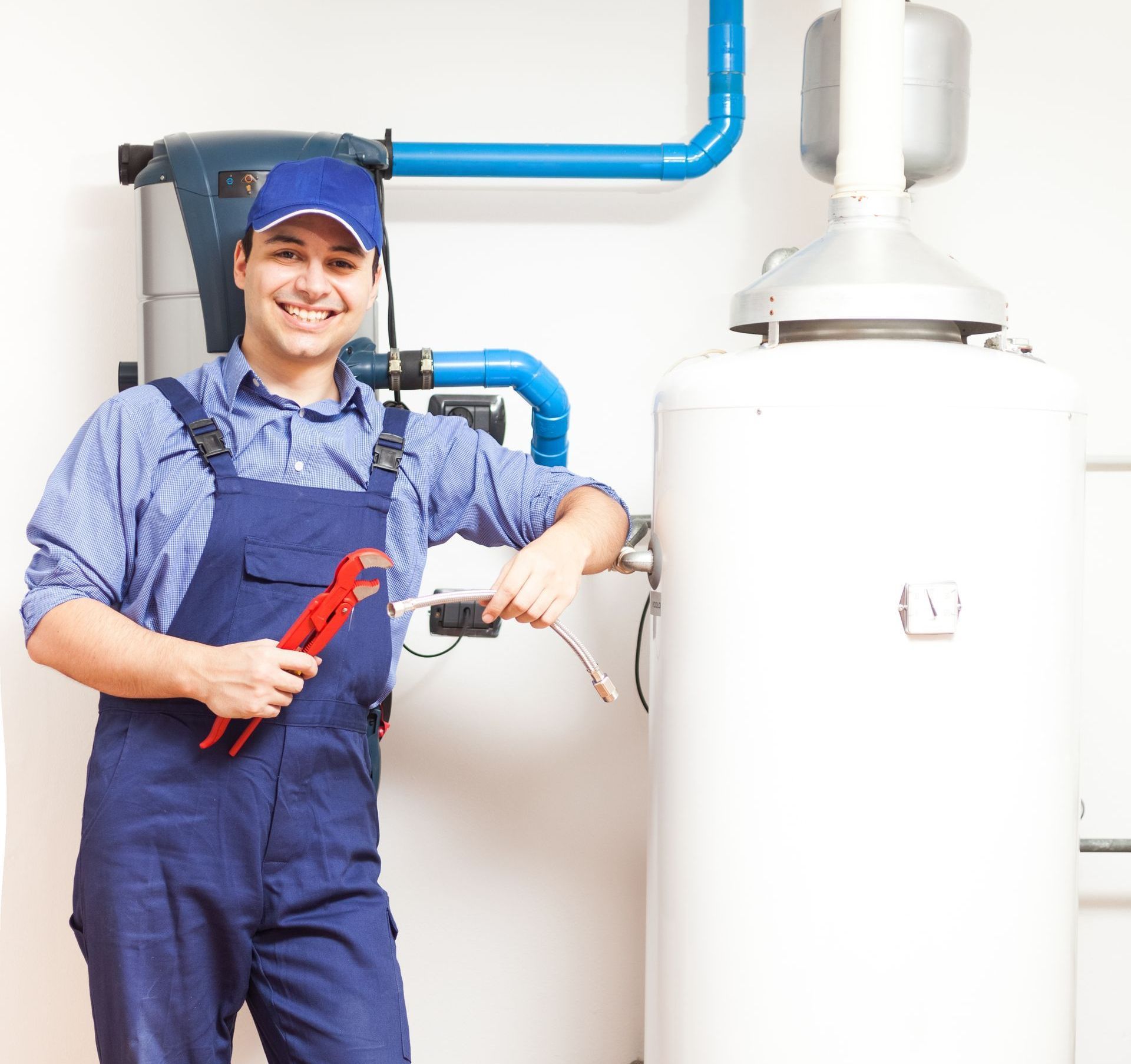 A man in blue overalls is holding a wrench in front of a water heater