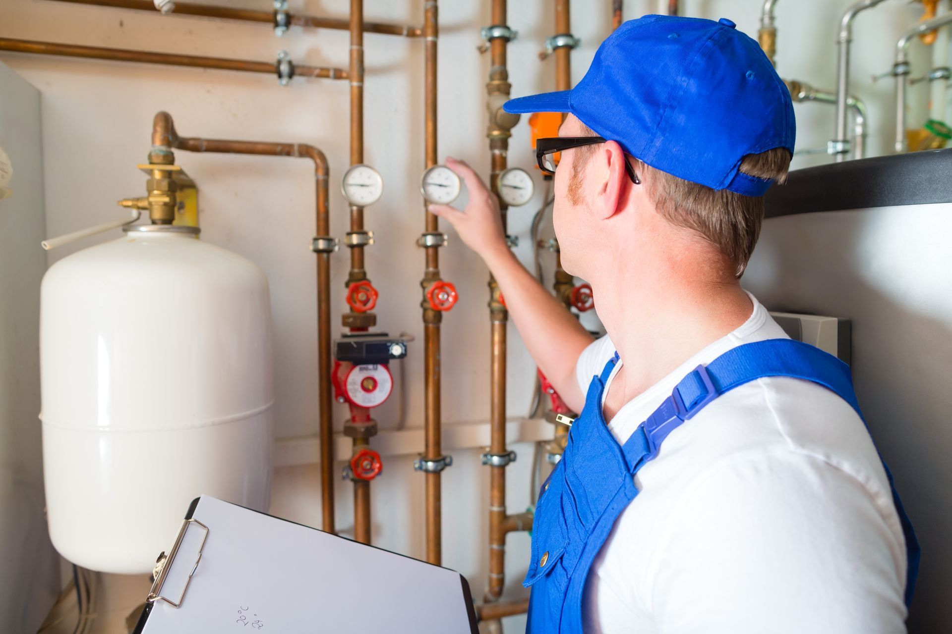 A man in a blue hat is working on a pipe while holding a clipboard.
