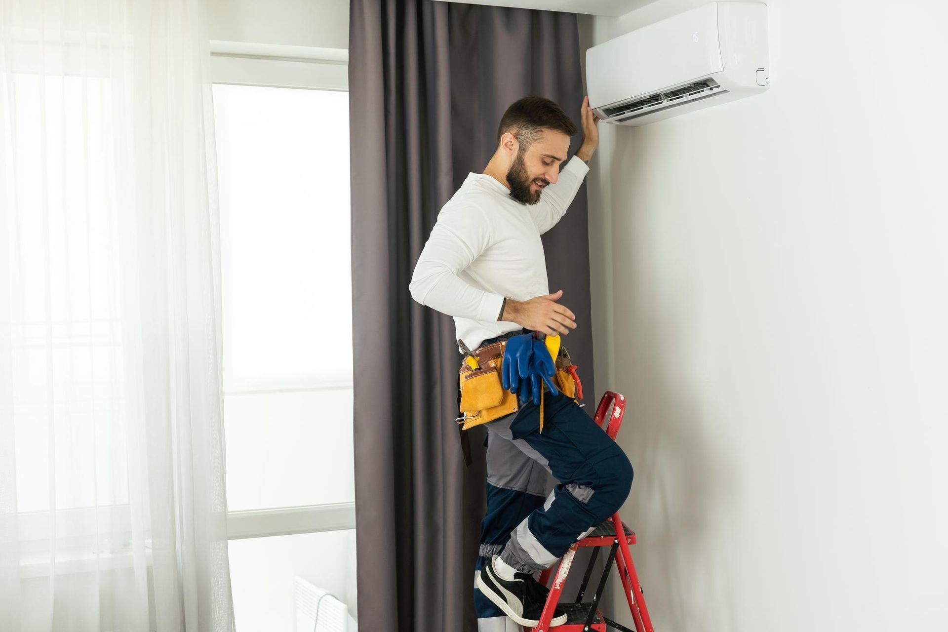 A man is standing on a ladder fixing an air conditioner.