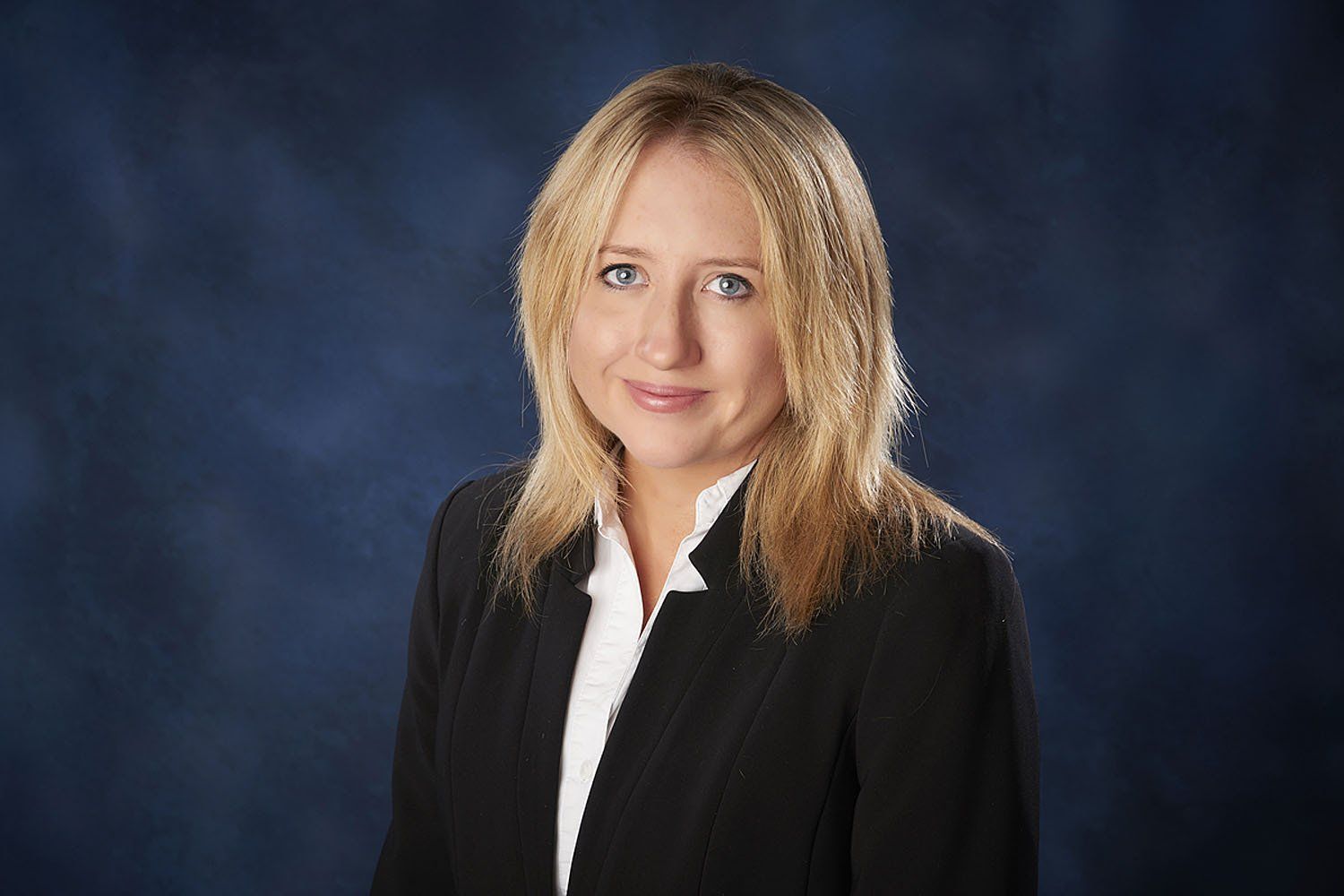 A woman in a black suit and white shirt is standing in front of a blue background.
