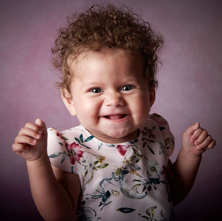 A baby with curly hair is wearing a floral dress and smiling