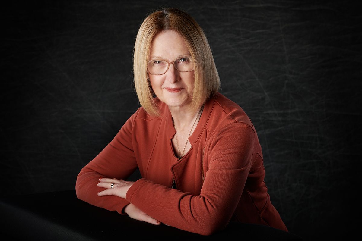 A woman wearing glasses and a red sweater is sitting at a table with her arms crossed.