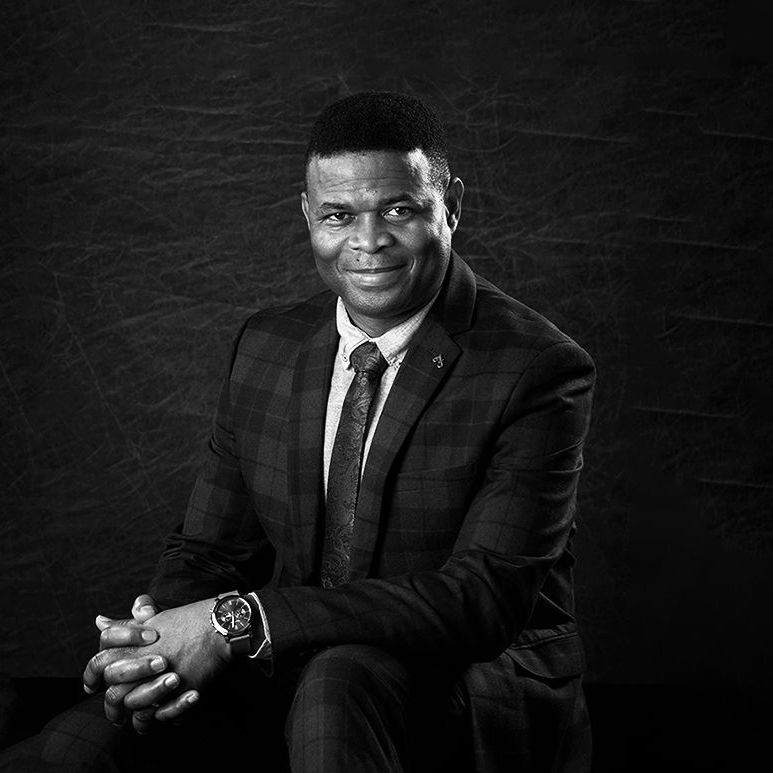 A black and white photo of a man in a suit and tie sitting with his hands folded.