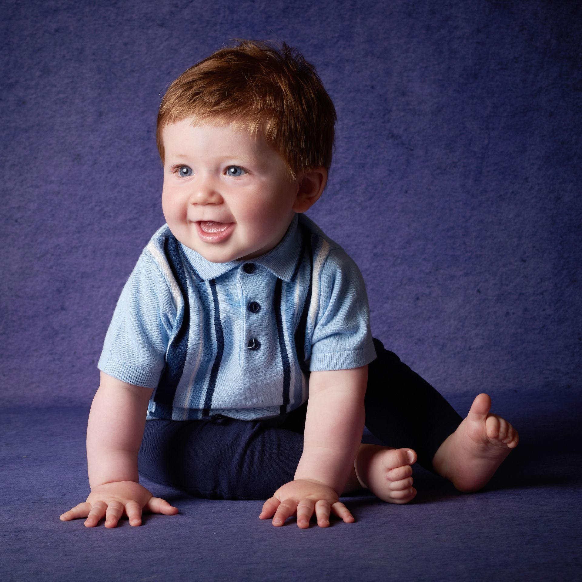 A baby in a blue shirt and suspenders is sitting on the floor