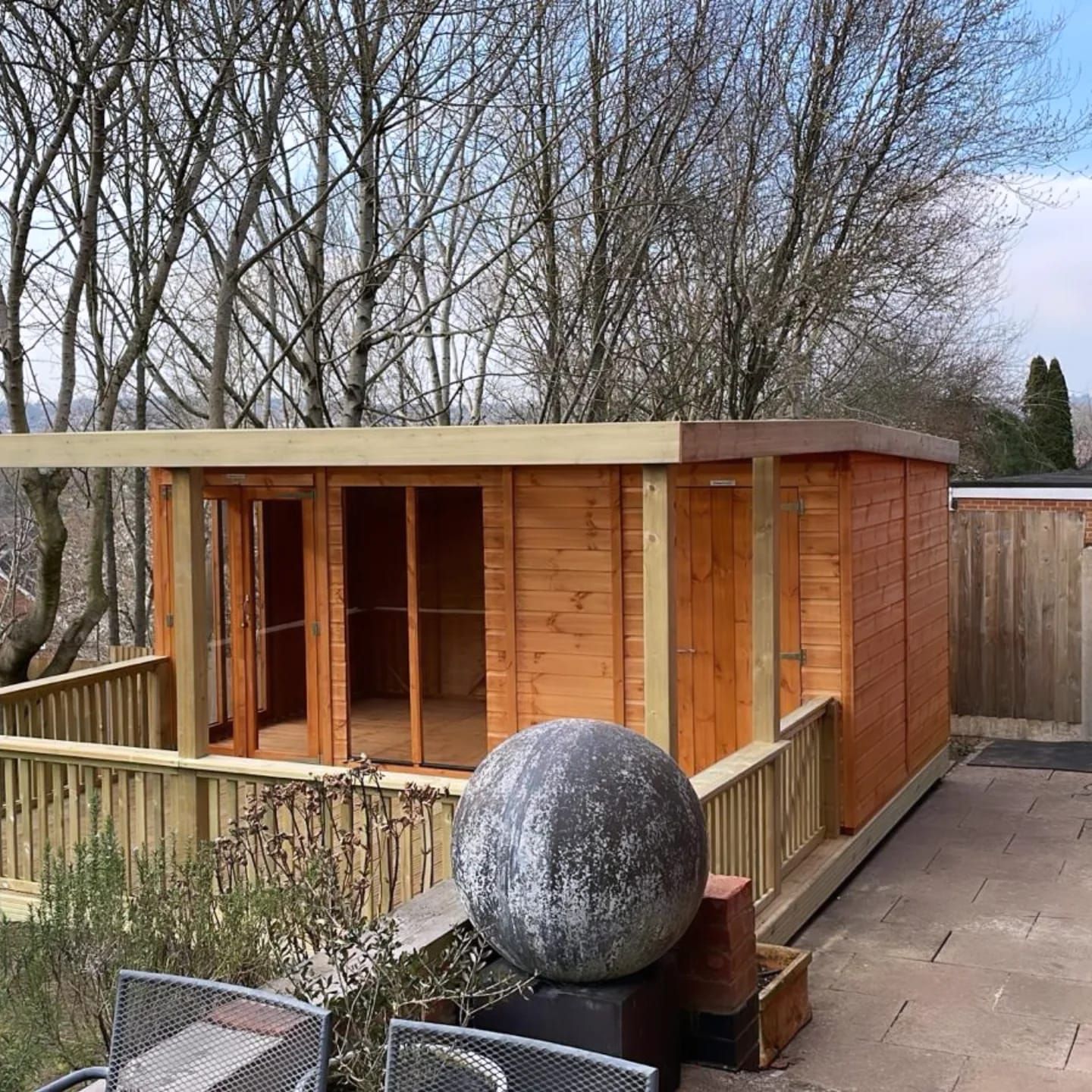 Wooden cabin with a wrap-around deck, set in a yard. A large stone sphere and outdoor furniture are nearby.