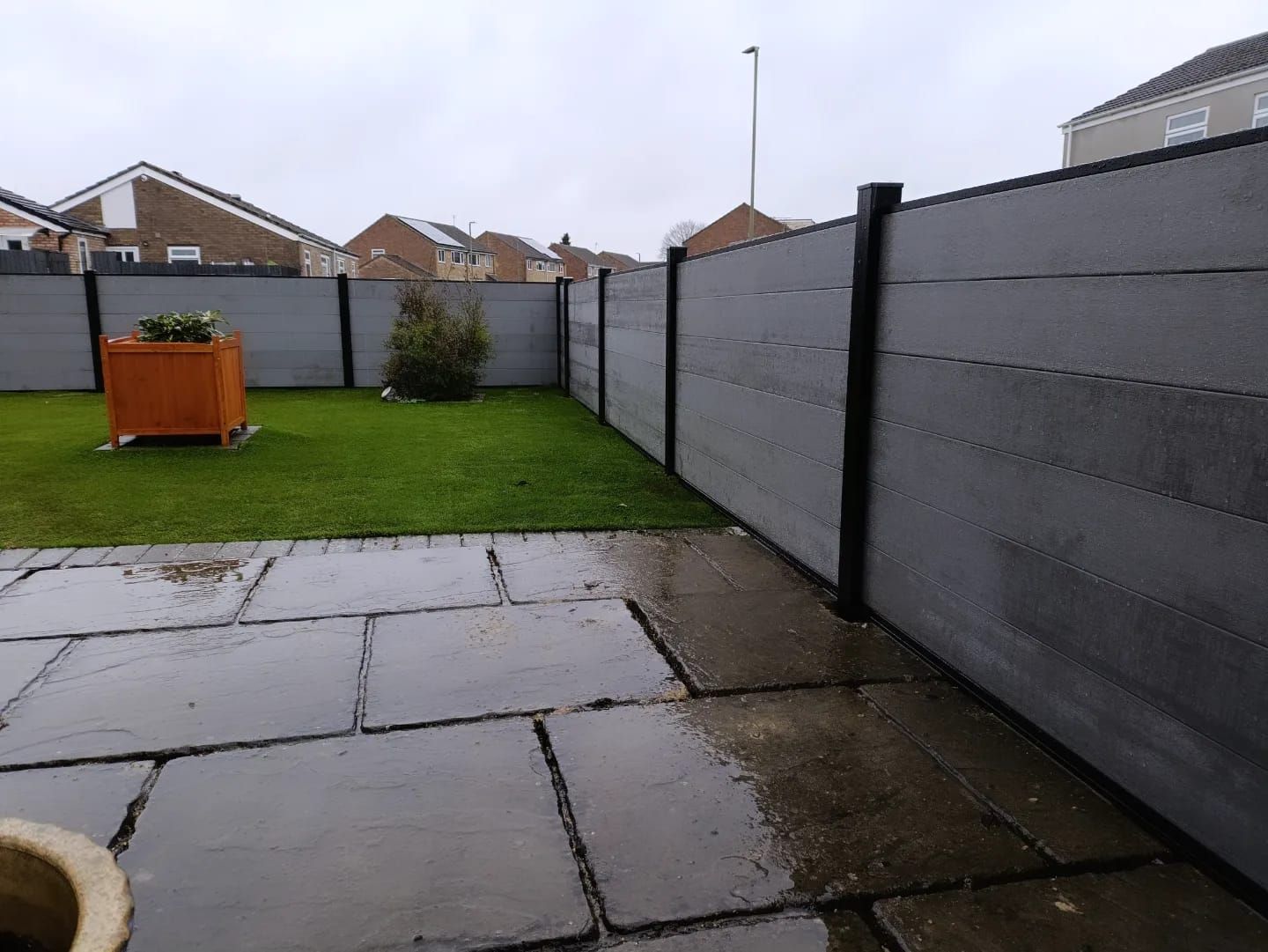 Gray composite fence in a backyard, with a paved patio in the foreground and grass. Overcast sky.