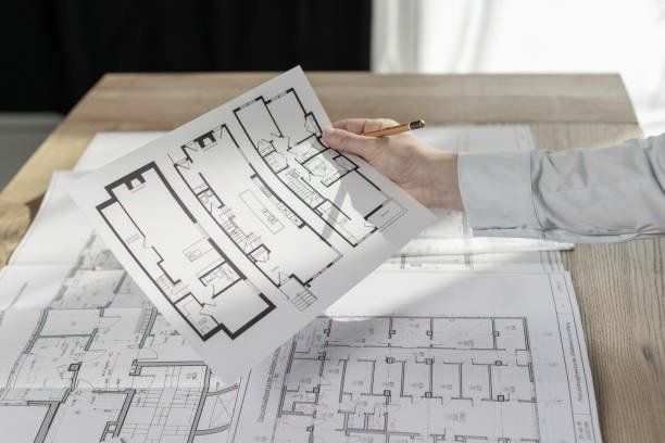 Person holding architectural floor plan over a wooden table with other blueprints.