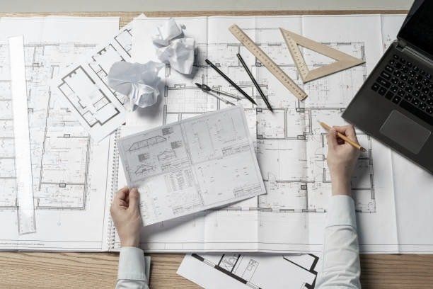 Architect at work on a blueprint, with tools and a laptop on a wooden table.