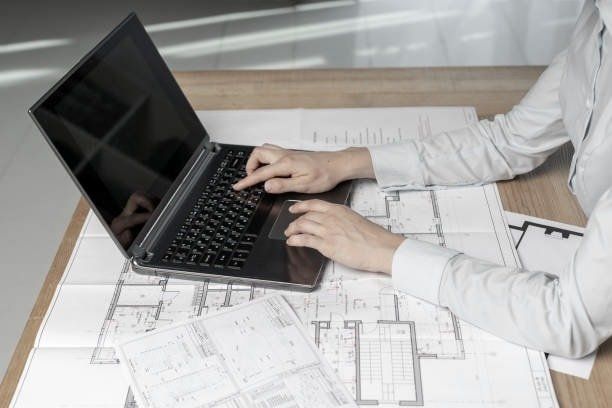 Person using laptop on a desk with blueprints; hands typing, office setting.