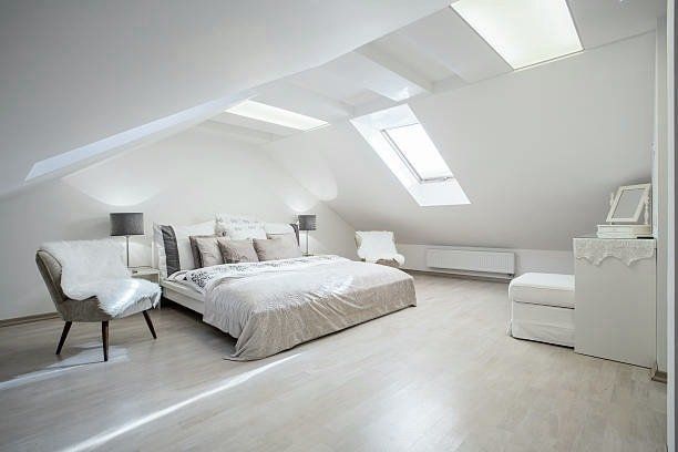 White bedroom with sloped ceiling, skylights, bed, chairs, dresser.