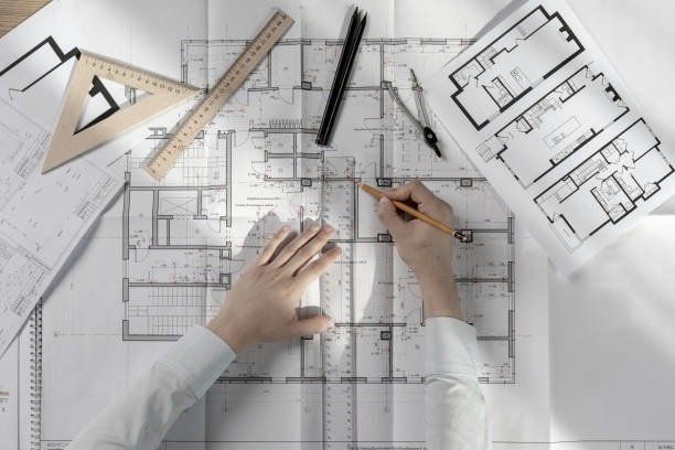 Hands drawing on blueprints with a pencil, surrounded by drafting tools.