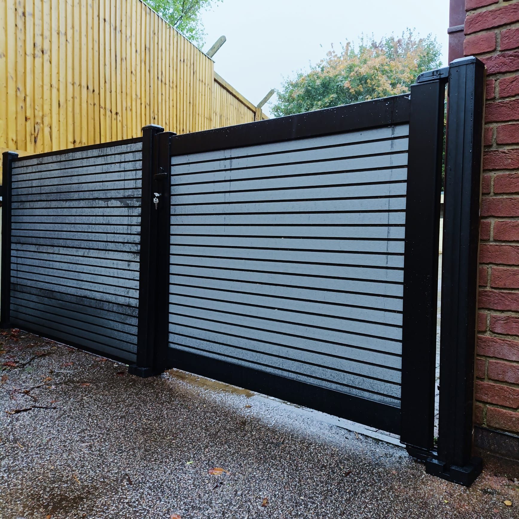 Black and gray slatted privacy gate with a wooden fence to the side, set on a concrete surface next to a brick wall.