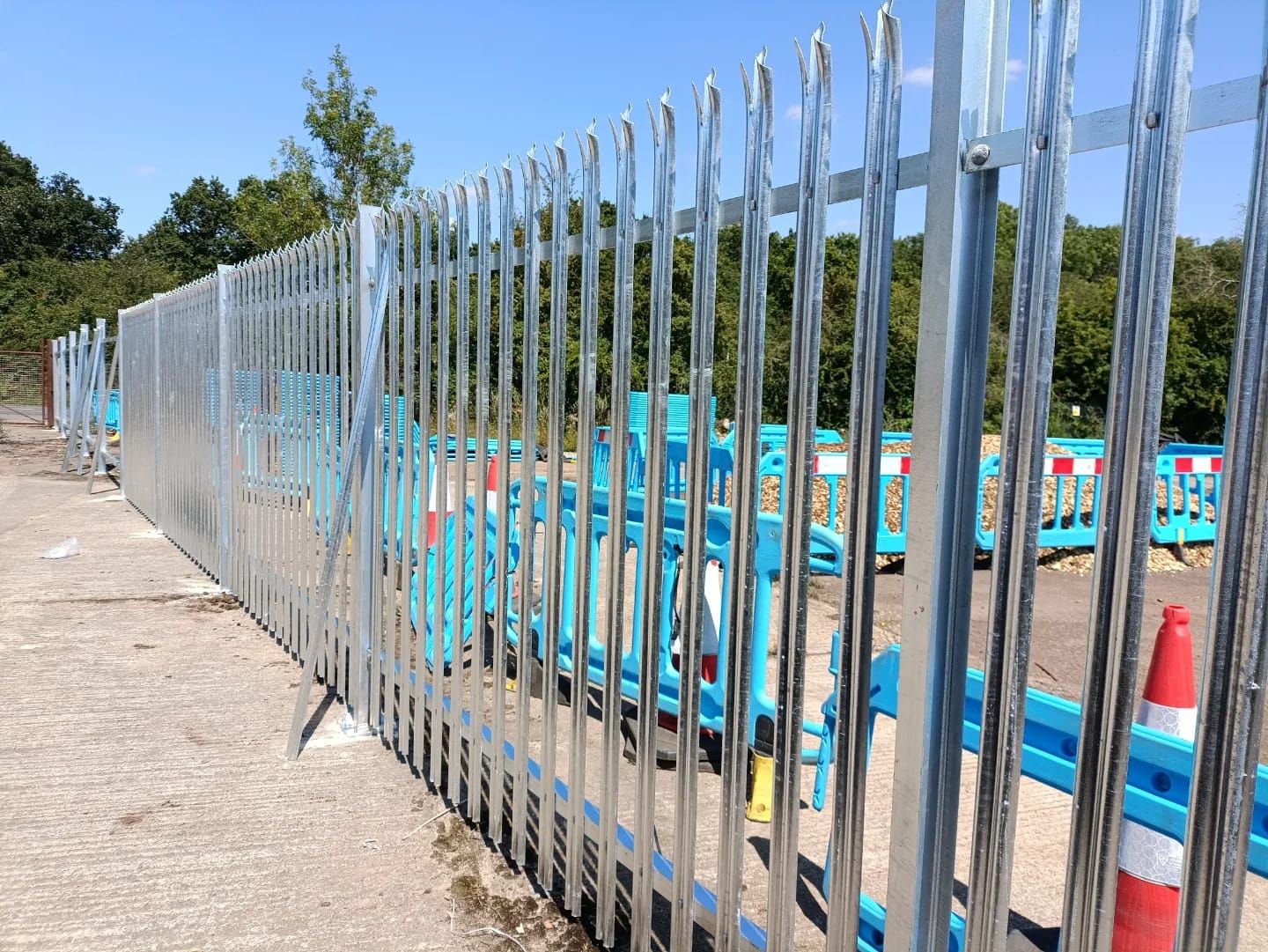 Galvanized steel fence. Construction zone with blue barriers and orange cone. Forest in the background.