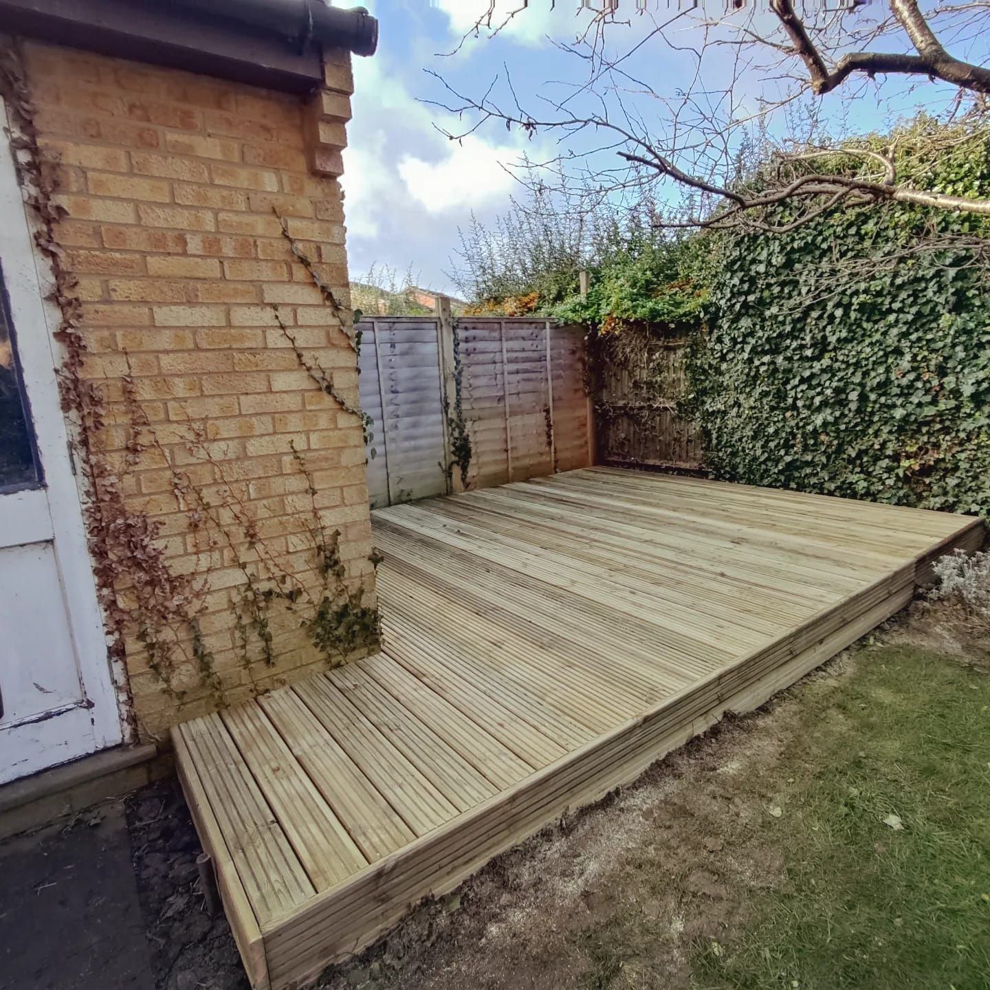 Wooden deck built in a backyard next to a brick wall, with ivy and a fence.