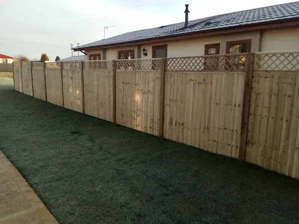 Wooden fence along a green lawn, bordering a beige house with a dark roof on a sunny day.