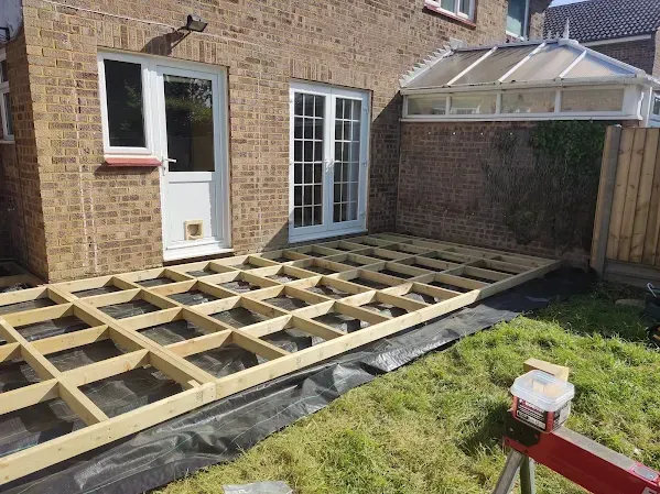 Wooden deck frame construction against a brick house, with grass and a small conservatory visible.