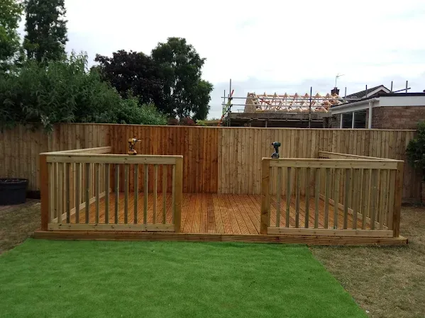 Wooden deck with railing in a backyard. Brown fence and green grass surround it.