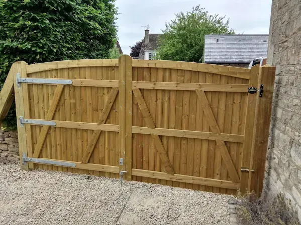 Wooden double gate, light brown color, set in stone wall, opens onto gravel driveway.