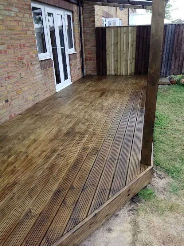 Wooden deck with a covered section against a brick building and fence; a grassy lawn borders it.