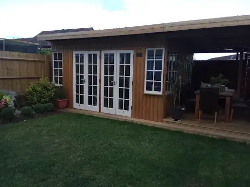 Wooden shed with French doors and a deck in a backyard with green grass and a fence.