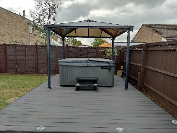 A gray hot tub on a deck under a gazebo in a backyard, surrounded by a wooden fence.