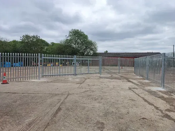 Metal security fence with a gate in a concrete lot, trees in the background under a cloudy sky.