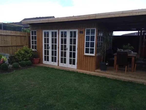 Wooden garden shed with French doors and windows, lawn in the foreground, fence and deck visible.