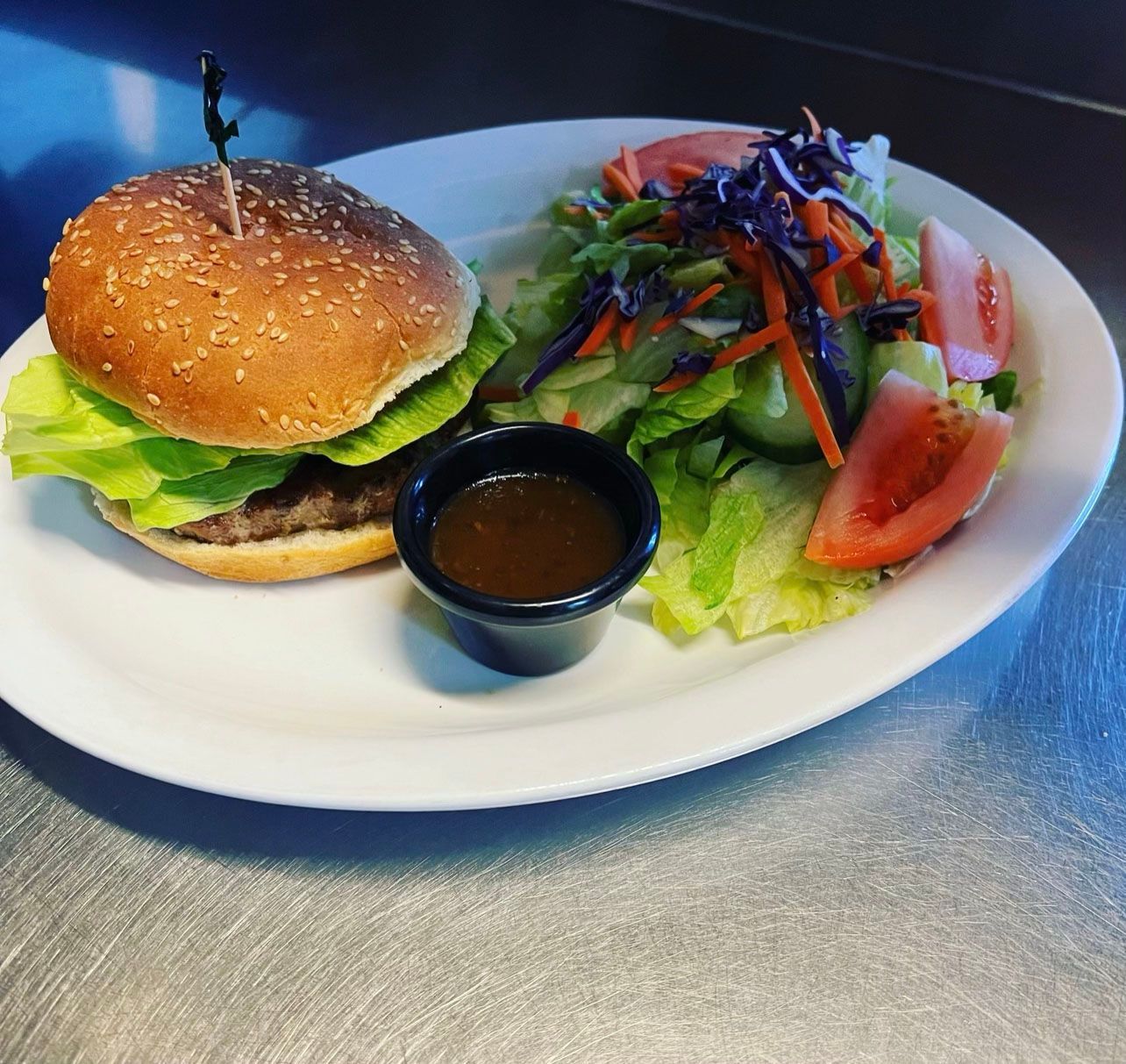 Burger with sesame bun, lettuce, and side salad with dressing on a white plate.