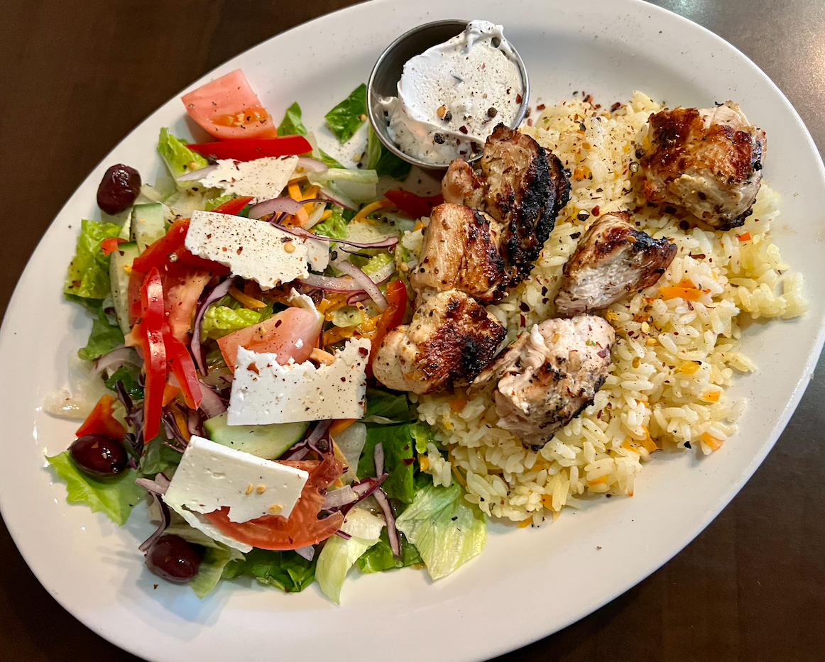 Grilled chicken skewers, Greek salad with feta, rice pilaf, and tzatziki on a white plate.