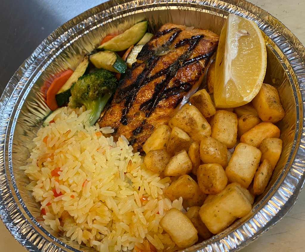 Grilled salmon with rice, roasted potatoes, vegetables, and a lemon wedge in a foil container.