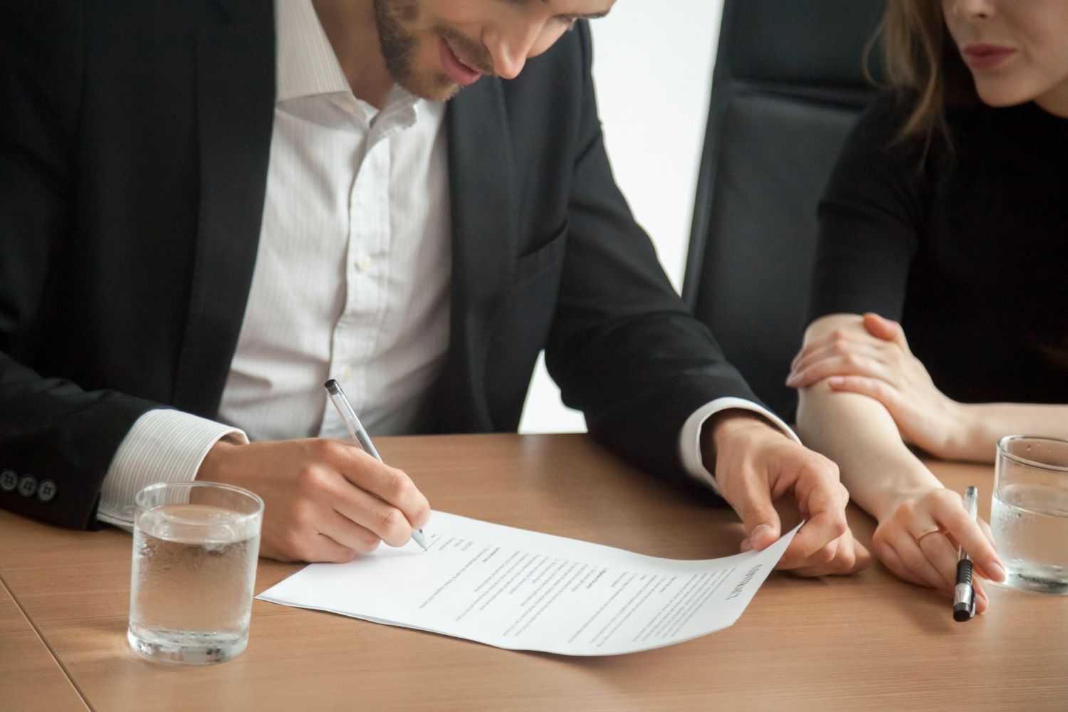 Two people in business attire review and sign a document at a wooden table with a glass of water.