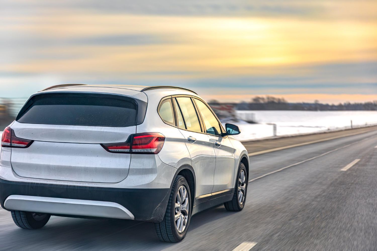 Silver SUV driving on an open road at sunset, with snowy fields and a blurred horizon.