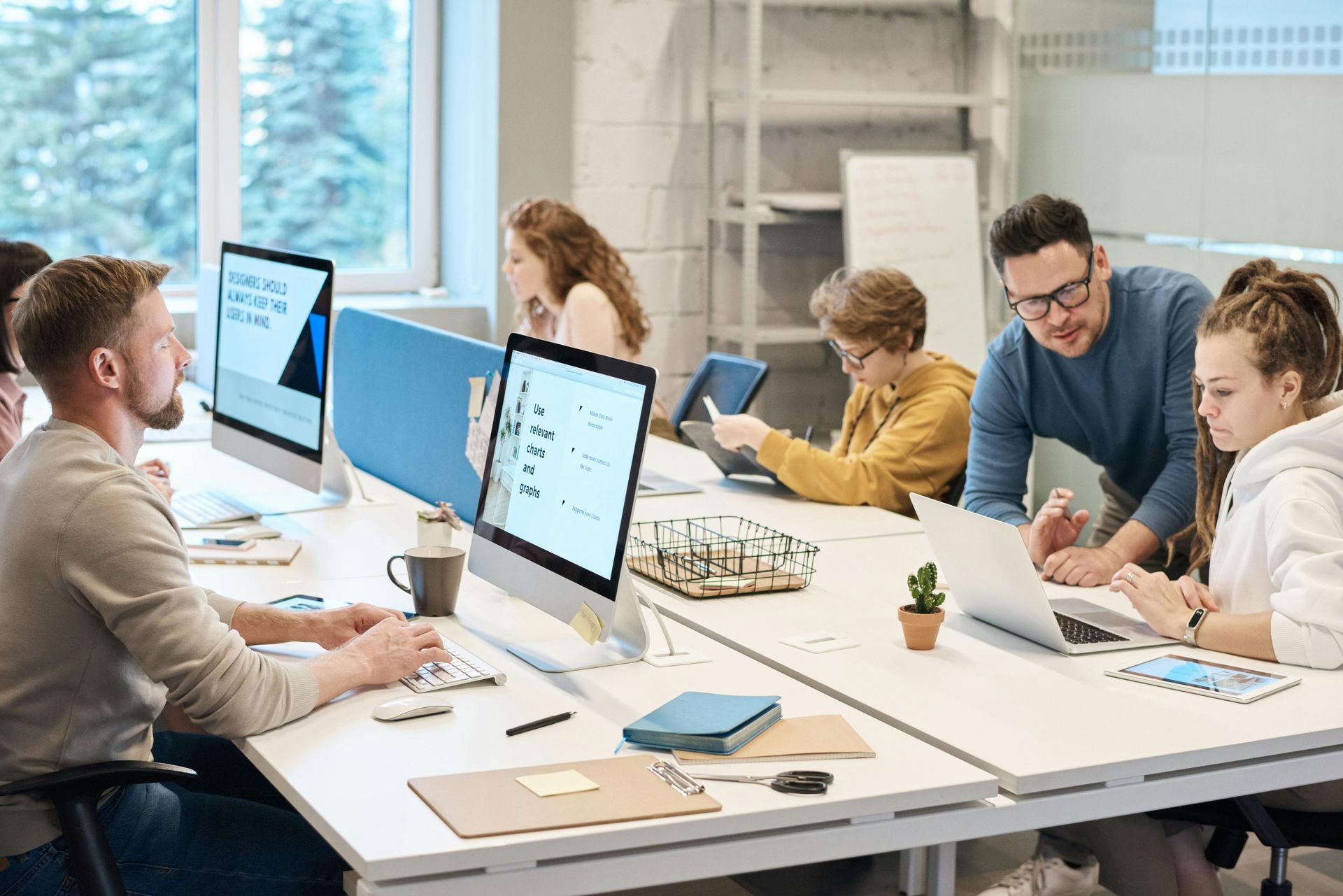 Team collaborating around desks in a bright office with computers, papers, and notebooks.