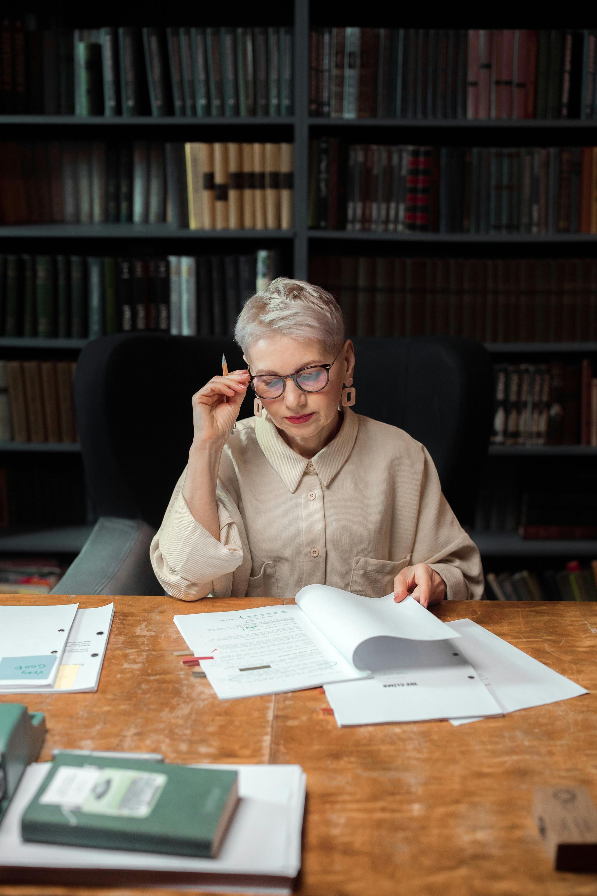 Person at a desk in a library, reading papers and adjusting glasses.