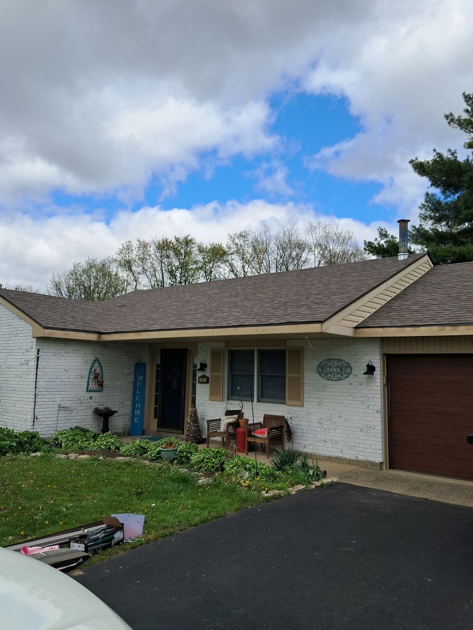 Single-story house with brown roof, white siding, black asphalt driveway, and a few chairs outside the front door.