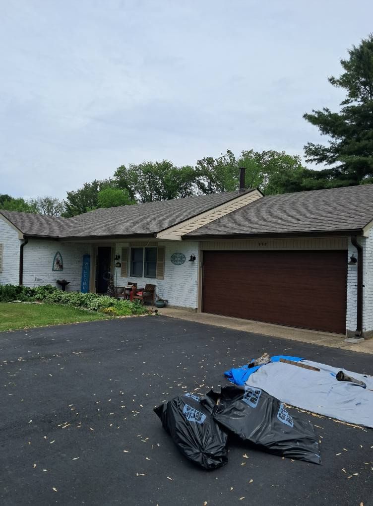 House with brown garage door, asphalt driveway. Two large black bags and tarp in foreground. Cloudy sky.