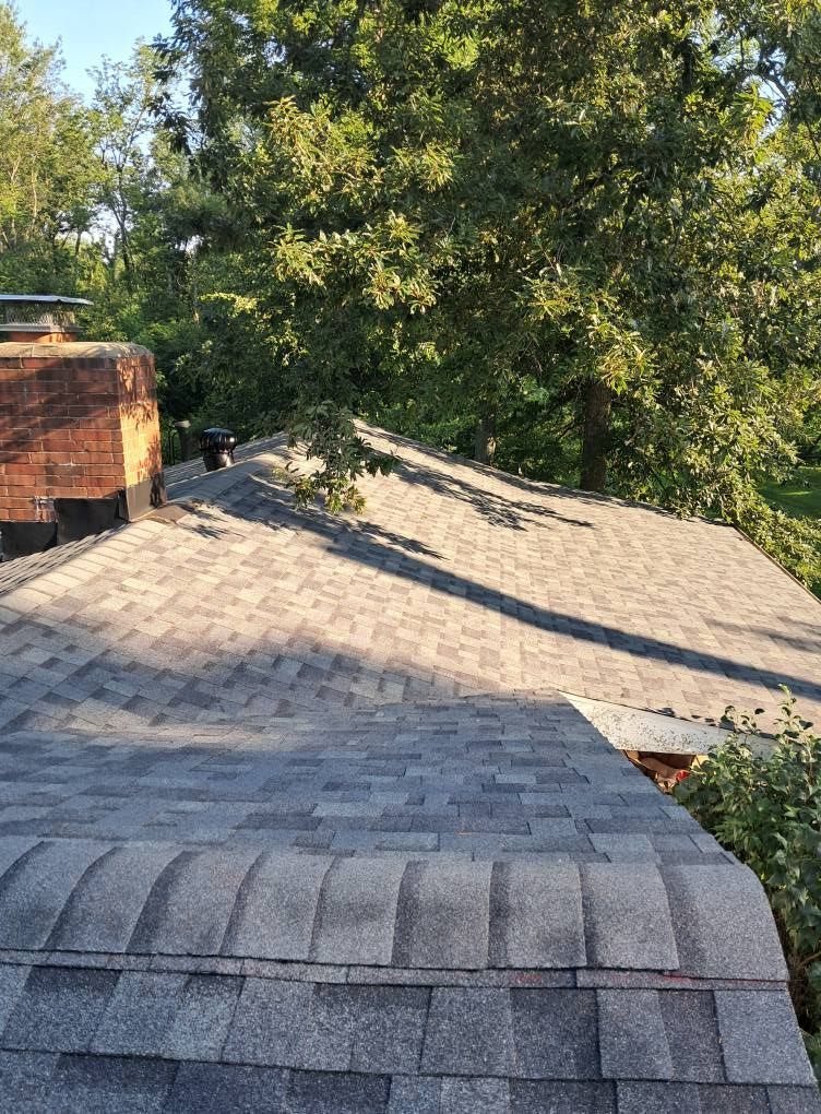 Asphalt shingle roof, gray with a brick chimney in the background, trees and blue sky.