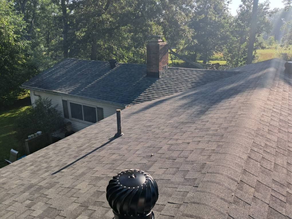 Shingled roofs of a house, with a chimney and trees in the background, under bright sunlight.